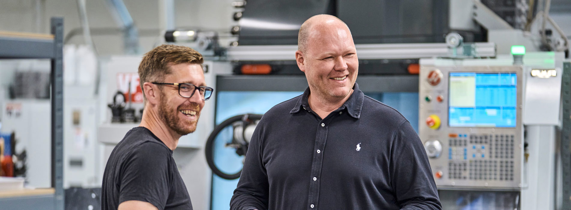 To kolleger fra Induflex står foran en drejebænk og snakker om et plastemne.