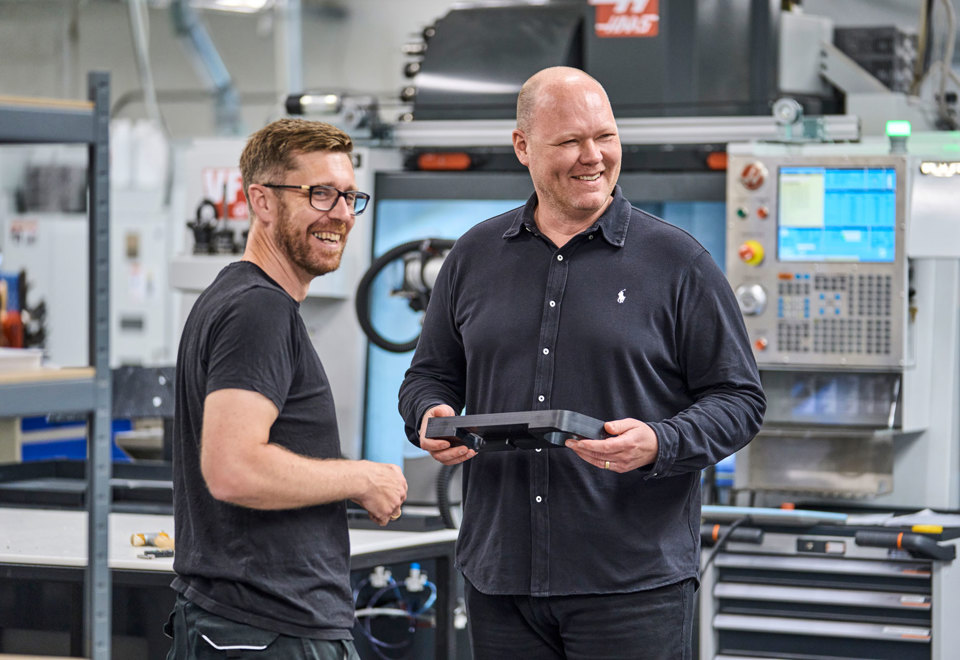 To kolleger fra Induflex står foran en drejebænk og snakker om et plastemne.