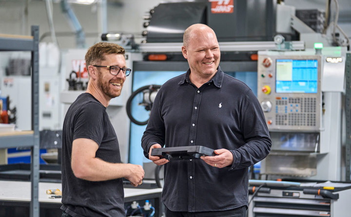 To kolleger fra Induflex står foran en drejebænk og snakker om et plastemne.