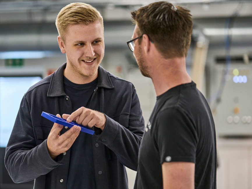 A salesperson points to a blue plastic part in front of an industrial technician.