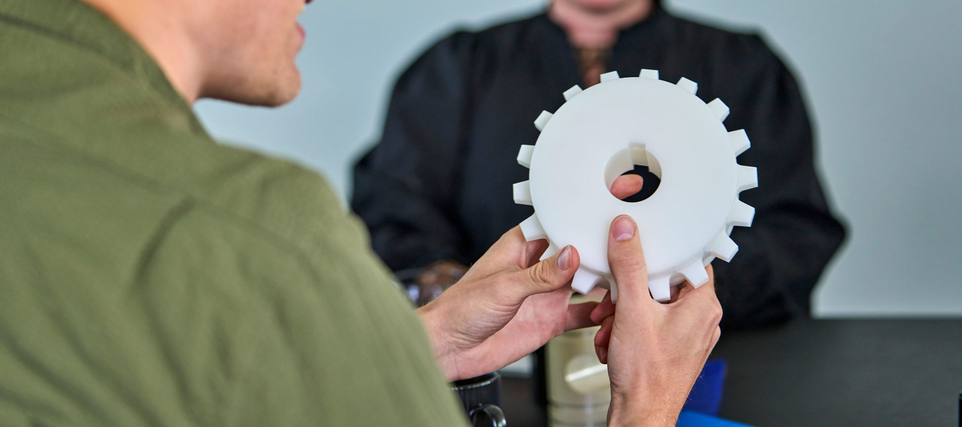 An employee holds a white machine part in his hands during a meeting.