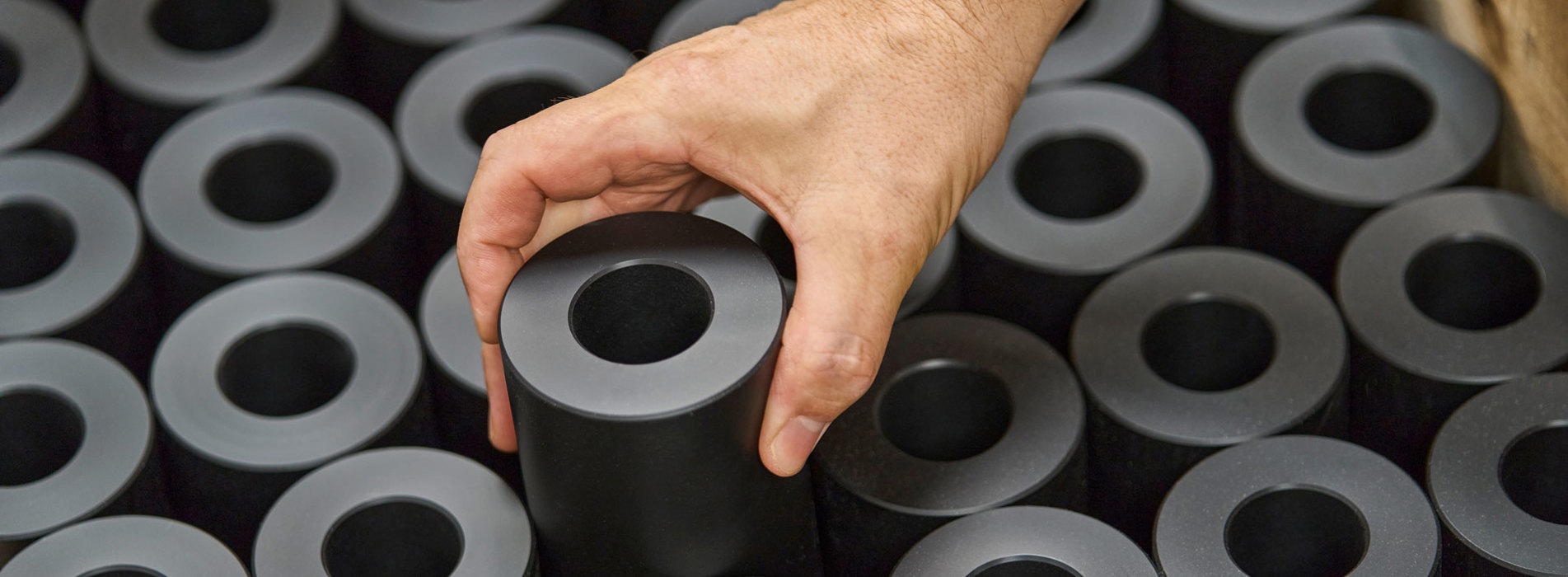 A hand lifts a turned machine part in black plastics from a pallet filled with machine parts.  