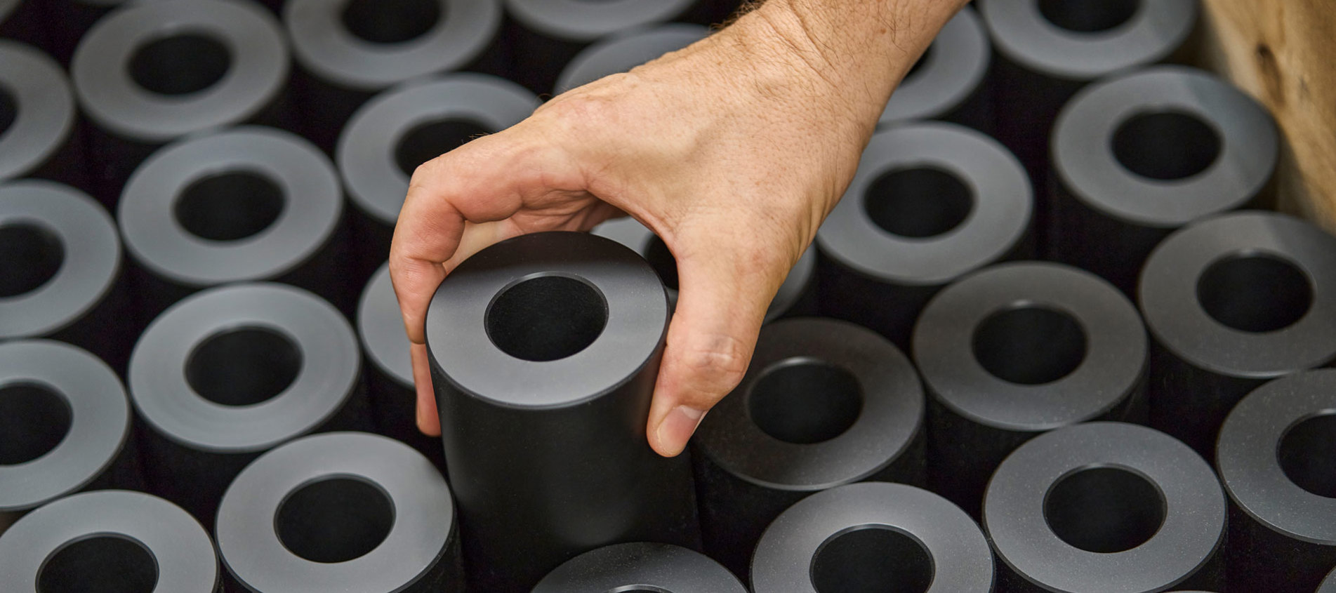 A hand lifts a turned machine part in black plastics from a pallet filled with machine parts.