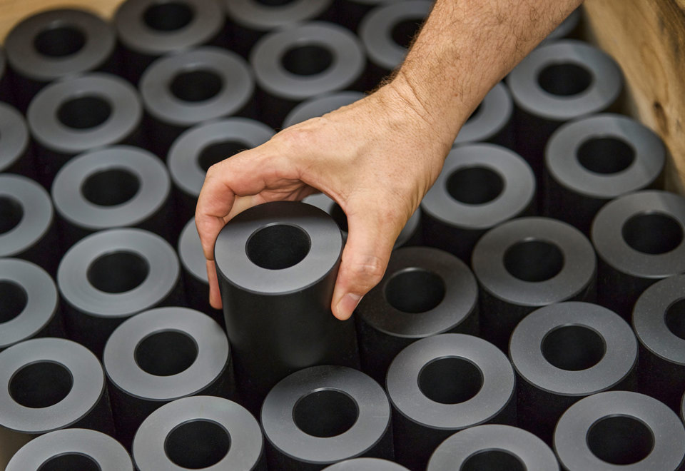 A hand lifts a turned machine part in black plastics from a pallet filled with machine parts.