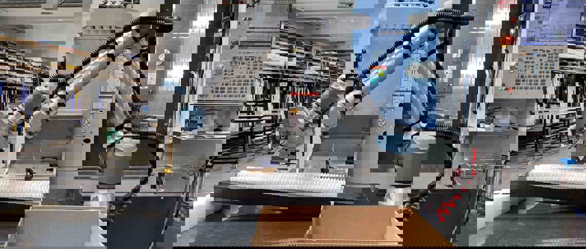 Robotarm plukker et hvidt plastemne fra et bord foran en CNC-fræser