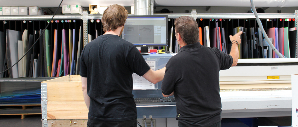 Two industry technician looking at the screen by a laser cutter, they are in the middle of preparing